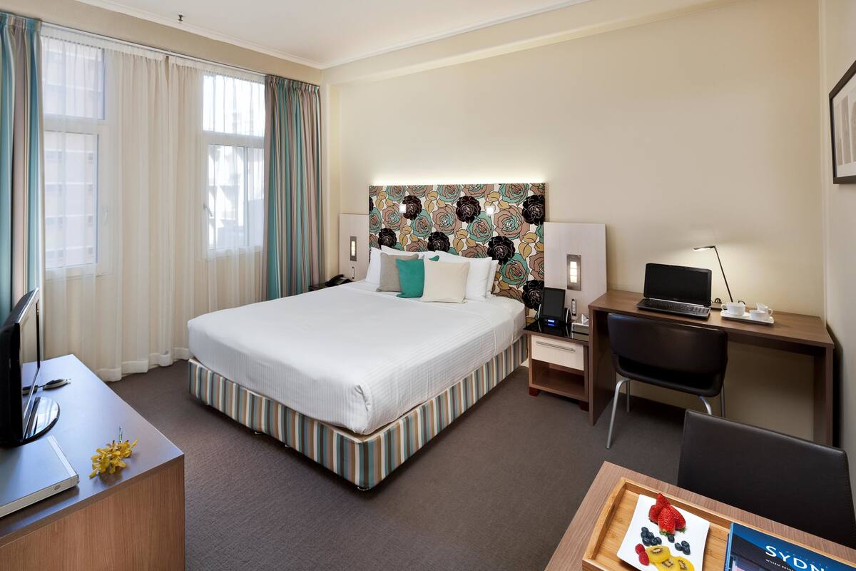 A hotel room with a large bed, a desk with a laptop, a chair, and a tray with fruit.