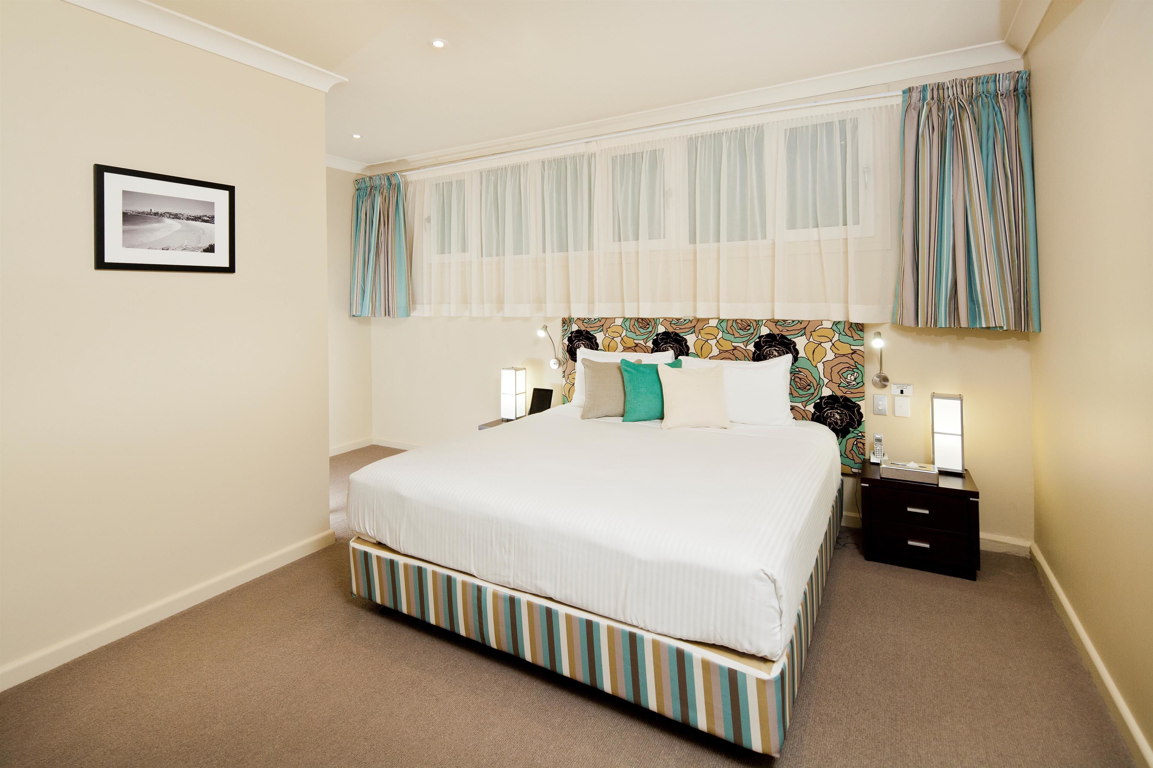 A neatly arranged bedroom with a large bed, a bedside table with a lamp, and a framed picture on the wall.