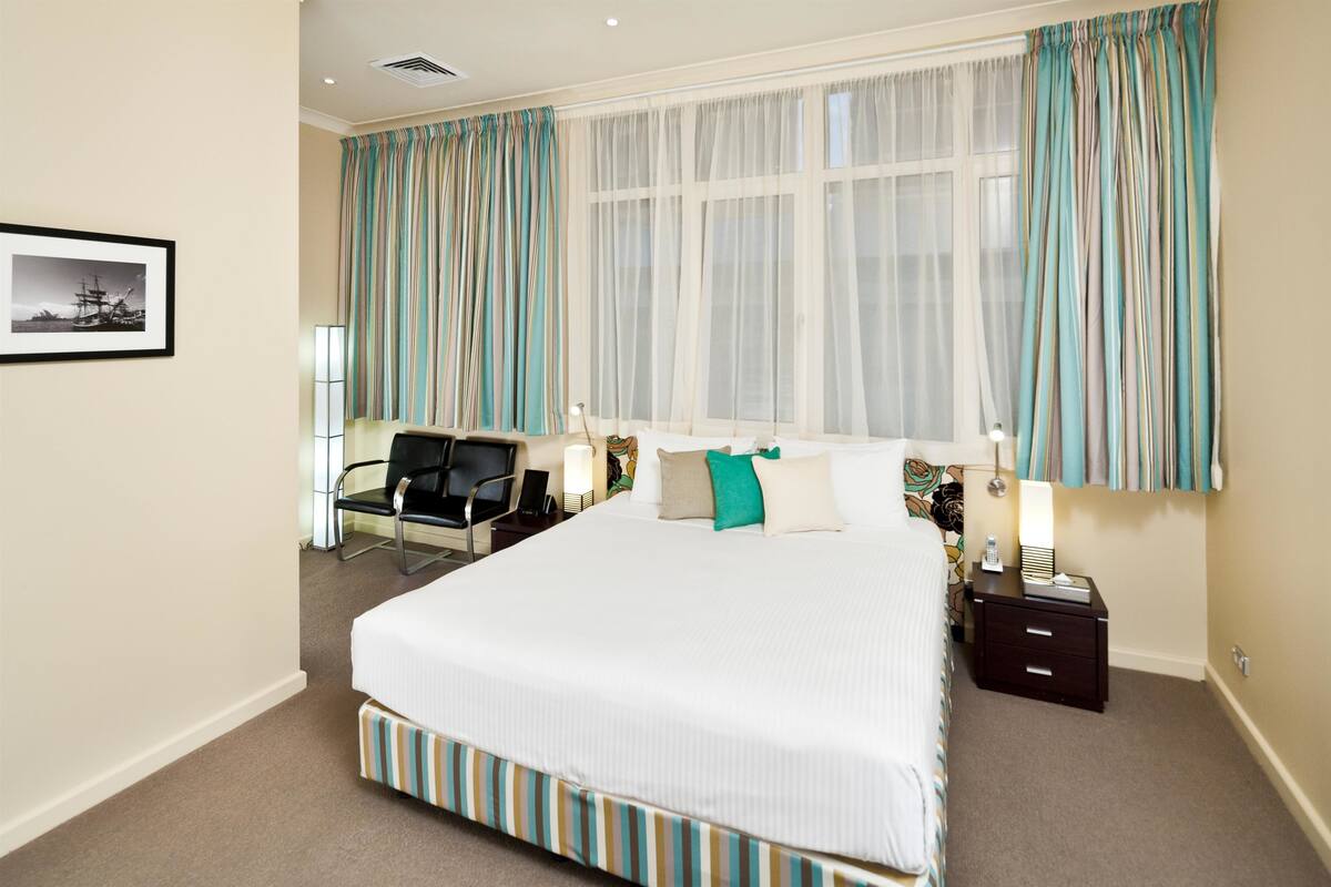 A hotel room with a large bed, a desk with a chair, a bedside table with a lamp, and a window with curtains.