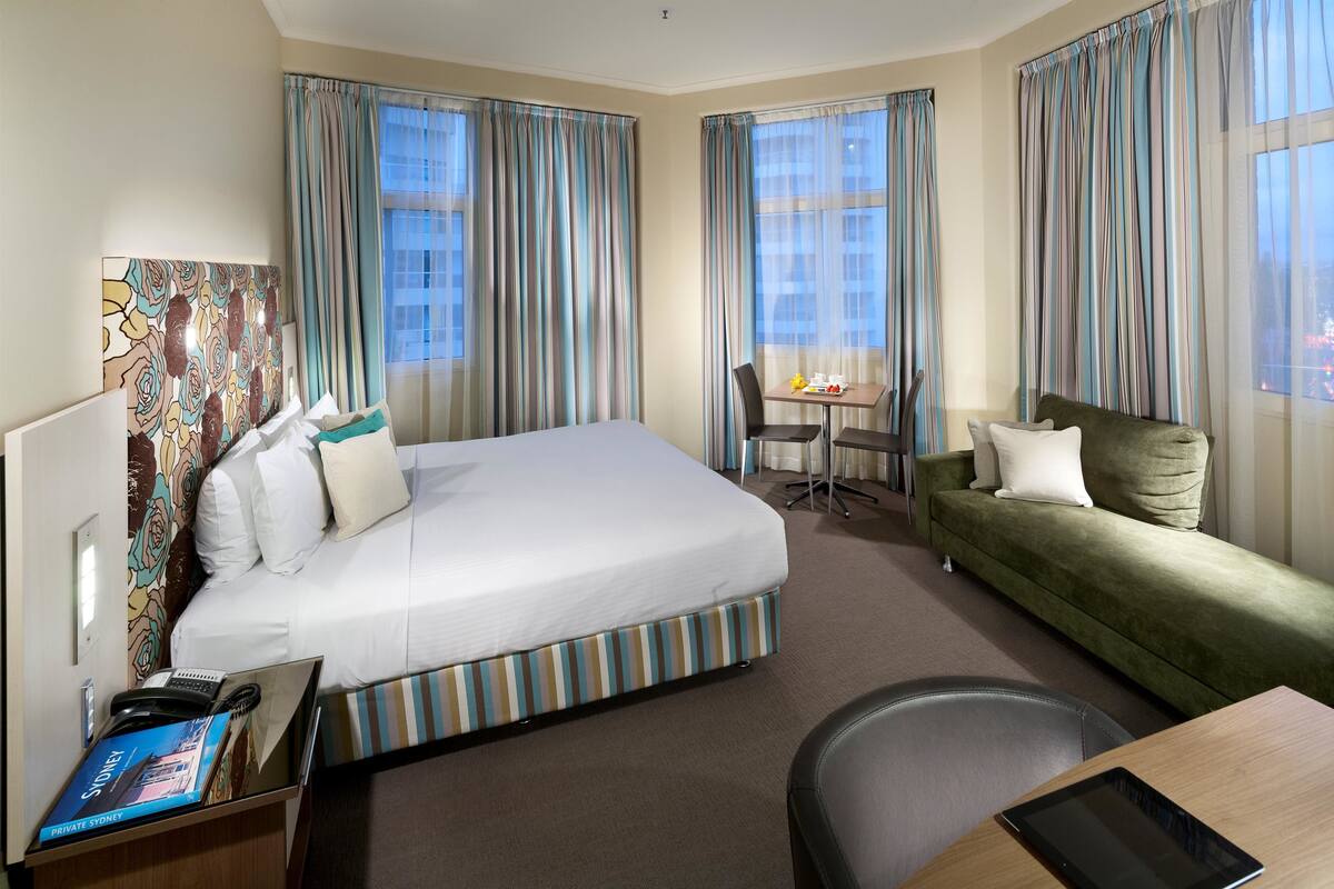 A hotel room with a large bed, a green sofa, a small table, and a chair.