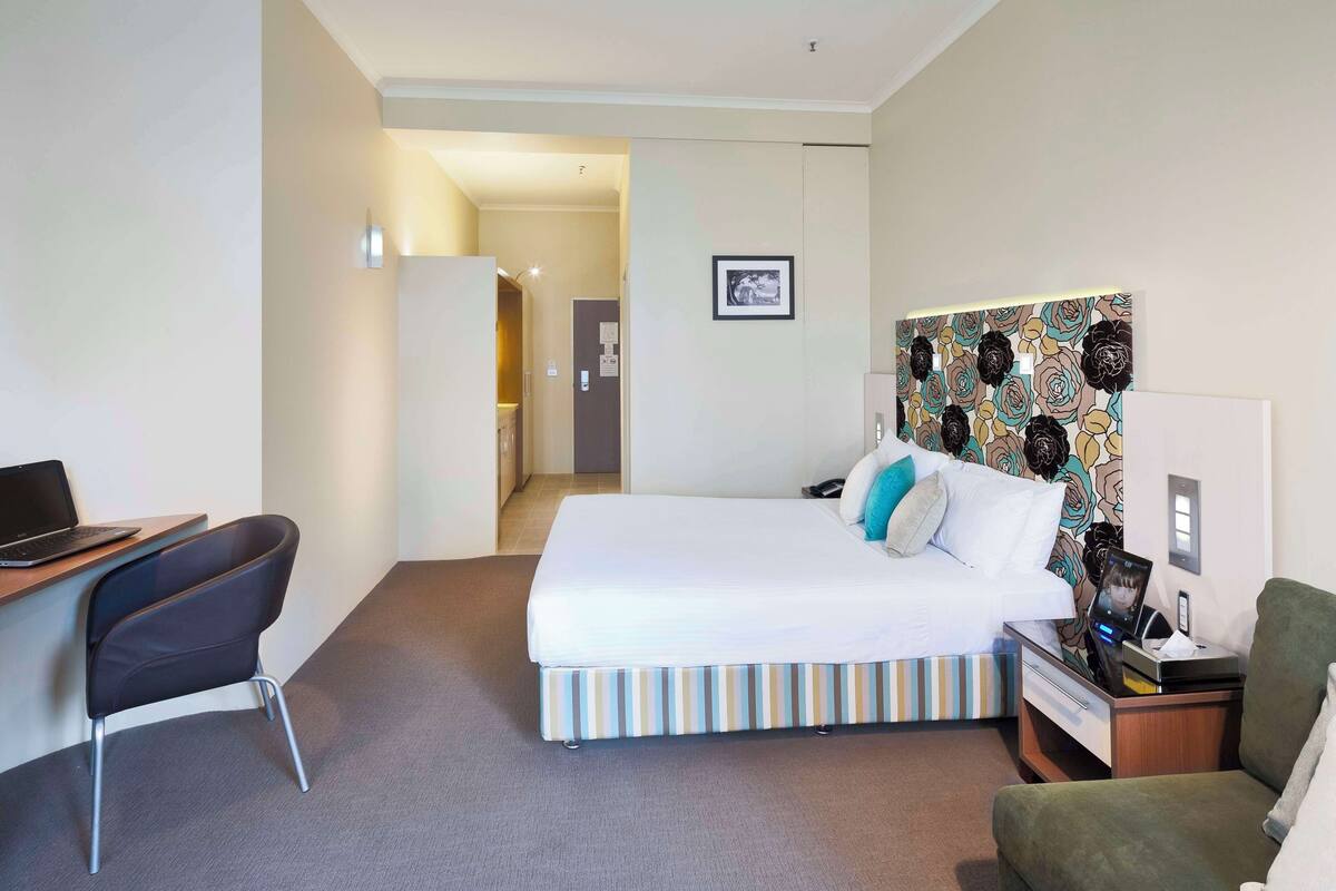A hotel room with a bed, a desk with a laptop, a chair, and a bedside table with a phone and a lamp.