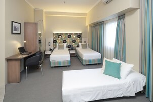 A hotel room with a desk, two single beds, a larger bed, a wardrobe, and a patterned headboard.