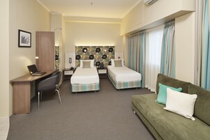 Una habitación de hotel con dos camas, un escritorio, un sofá verde y un cabecero estampado.