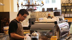Interior de una cafetería con un barista preparando bebidas, una máquina de café y una lámpara decorativa.