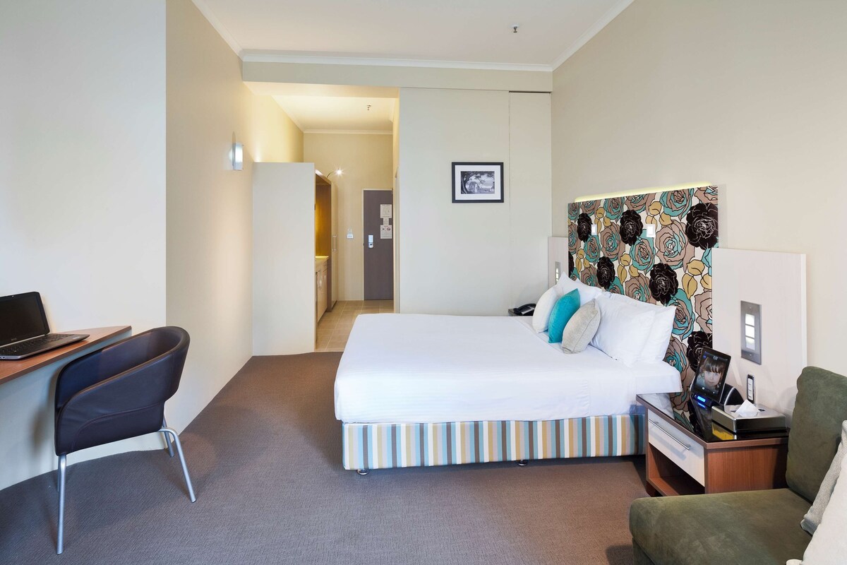 A hotel room with a bed, a desk with a laptop, a chair, and a small table with a phone and a lamp.