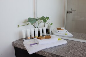 A bathroom with a mirror, a plant, and toiletries including body lotion, shampoo, and conditioner.