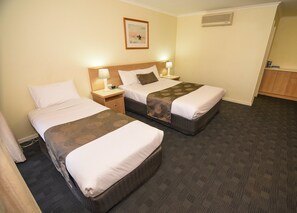 A hotel room with two beds, each with a pillow and a blanket, a nightstand with a lamp, a framed picture on the wall, and an air conditioning unit. - Sandown Heritage Motel (Noble Park)