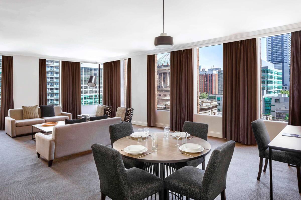 A modern hotel room with a dining area, a sofa set, and large windows offering city views.