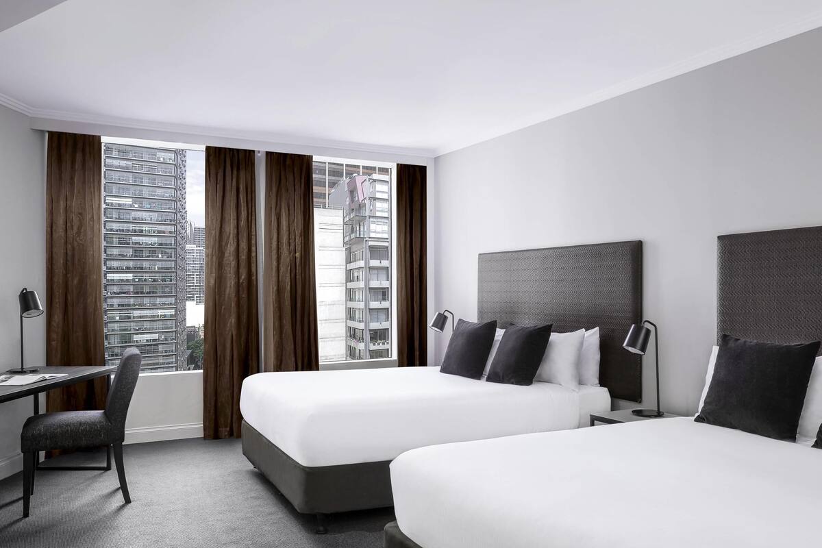 A hotel room with two beds, a desk, a chair, and a view of high-rise buildings.