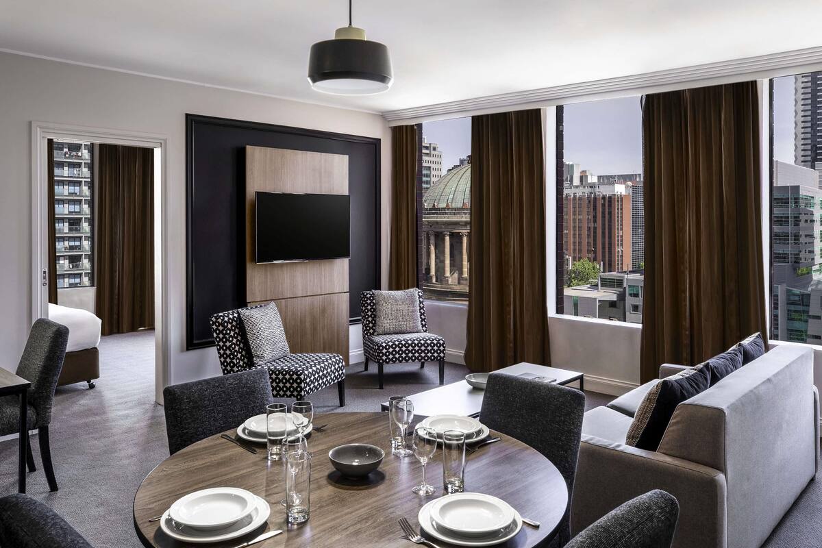 A modern hotel room with a dining area, a flat-screen TV mounted on a wooden panel, and a view of the cityscape through large windows.