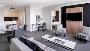 Un salon moderne comprenant un canapé, des fauteuils, une table basse, un coin repas avec une table et des chaises, un coin cuisine et un téléviseur fixé sur un panneau en bois.
