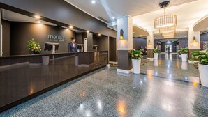 A modern hotel reception area with a dark reception desk, potted plants, and a spacious lobby with a polished floor.