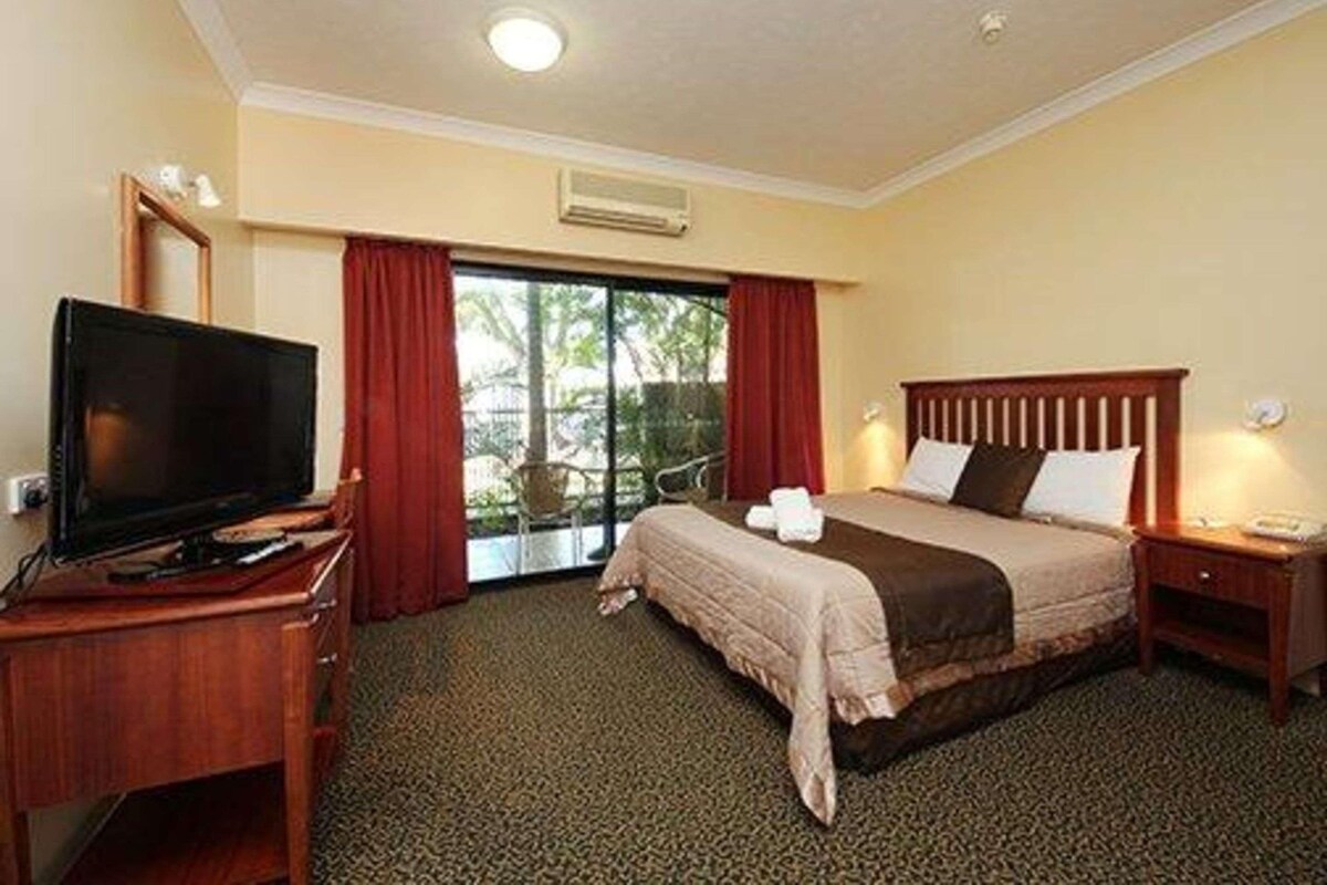 Una habitación de hotel con una cama, un televisor sobre un escritorio de madera y vistas a zonas verdes por la ventana.