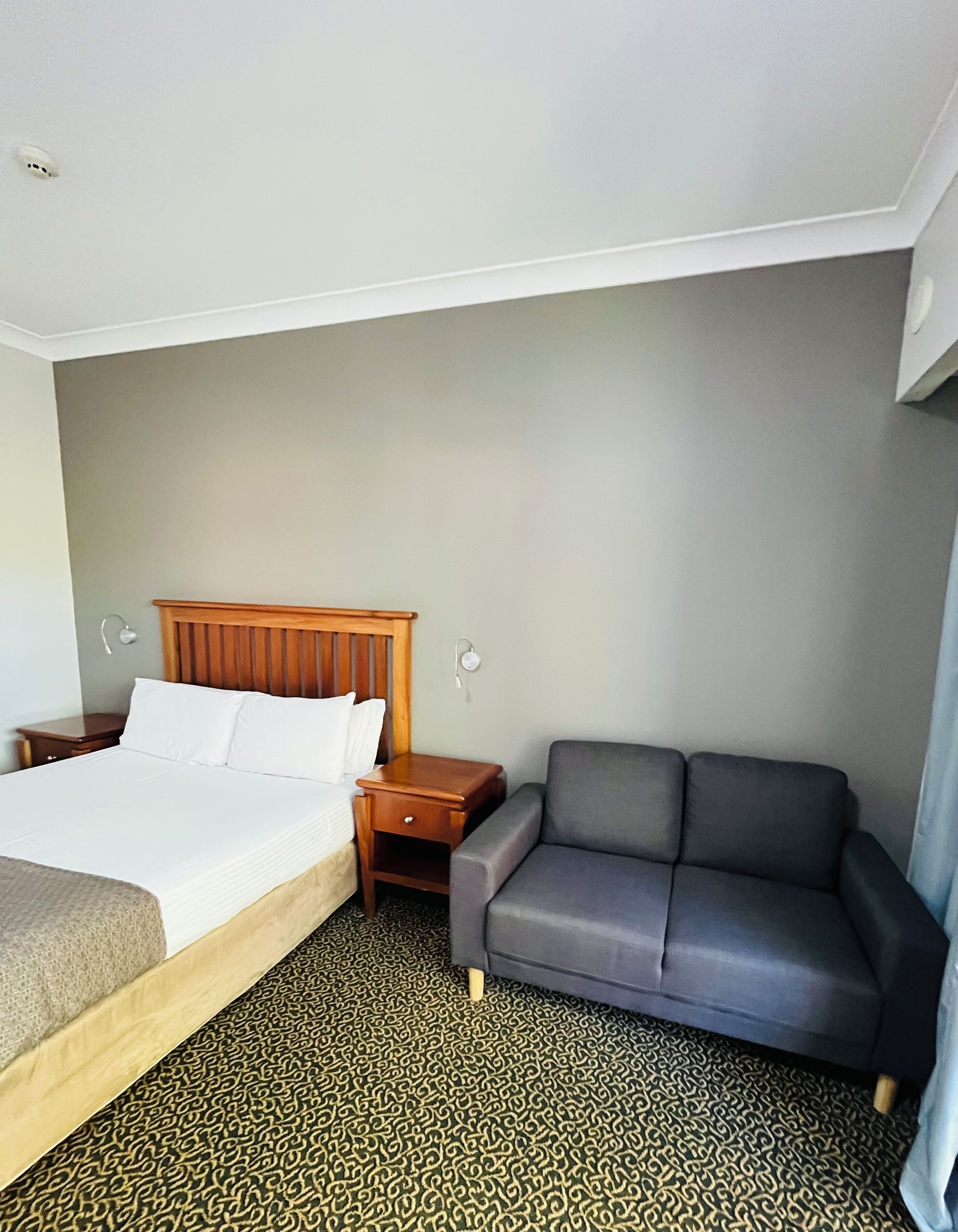 A hotel room with a bed, a wooden headboard, a grey sofa, a nightstand, and a patterned carpet.