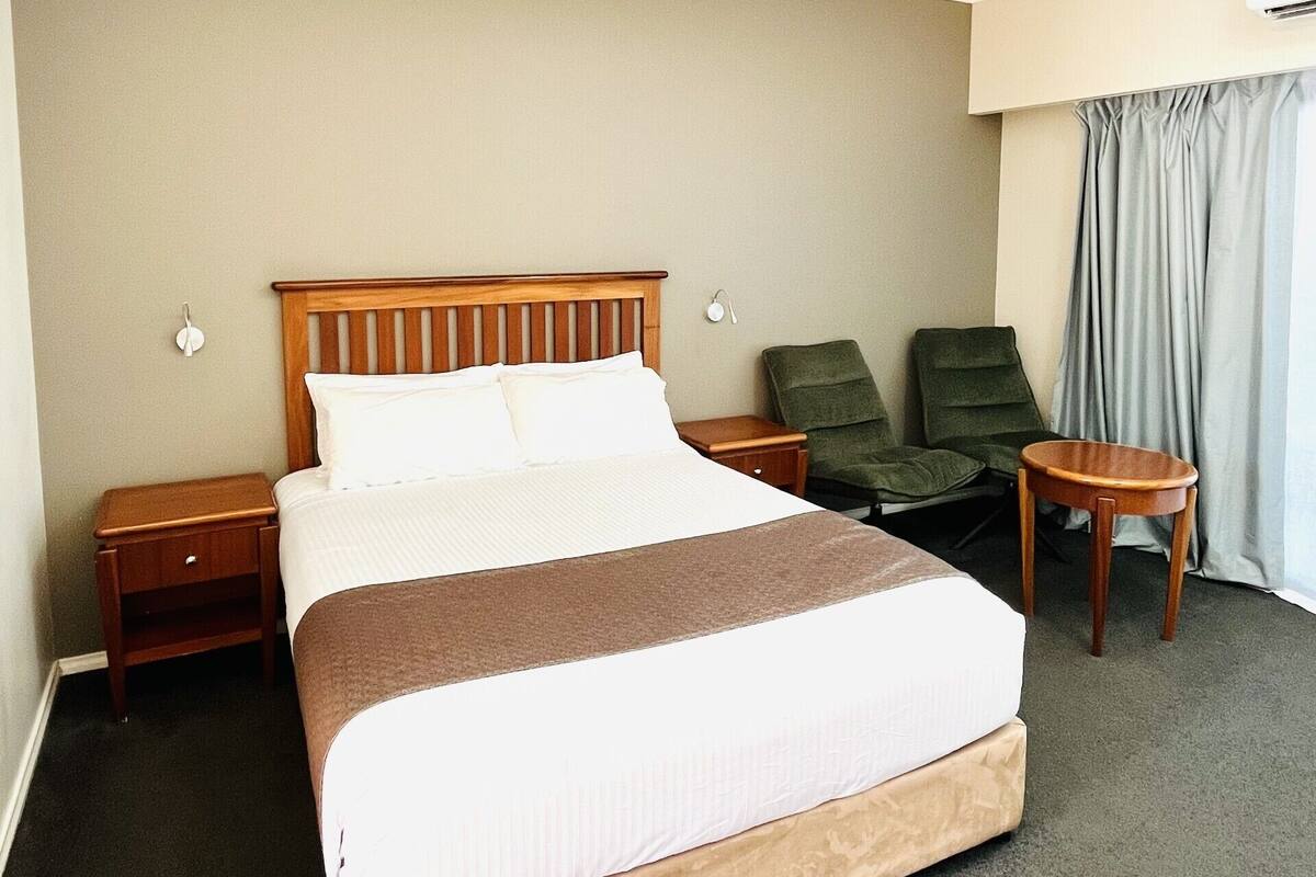 A hotel room with a large bed, two nightstands, a chair, a small table, and a window with curtains.