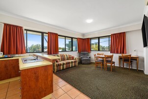 Una sala de estar moderna con sofá, una butaca, una mesa de comedor con sillas, una zona de cocina con fregadero y ventanas amplias con cortinas.