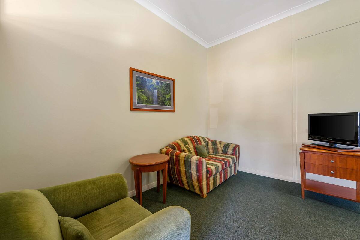 A small, sparsely furnished room with a green sofa, a striped armchair, a small wooden table, and a television on a wooden stand.