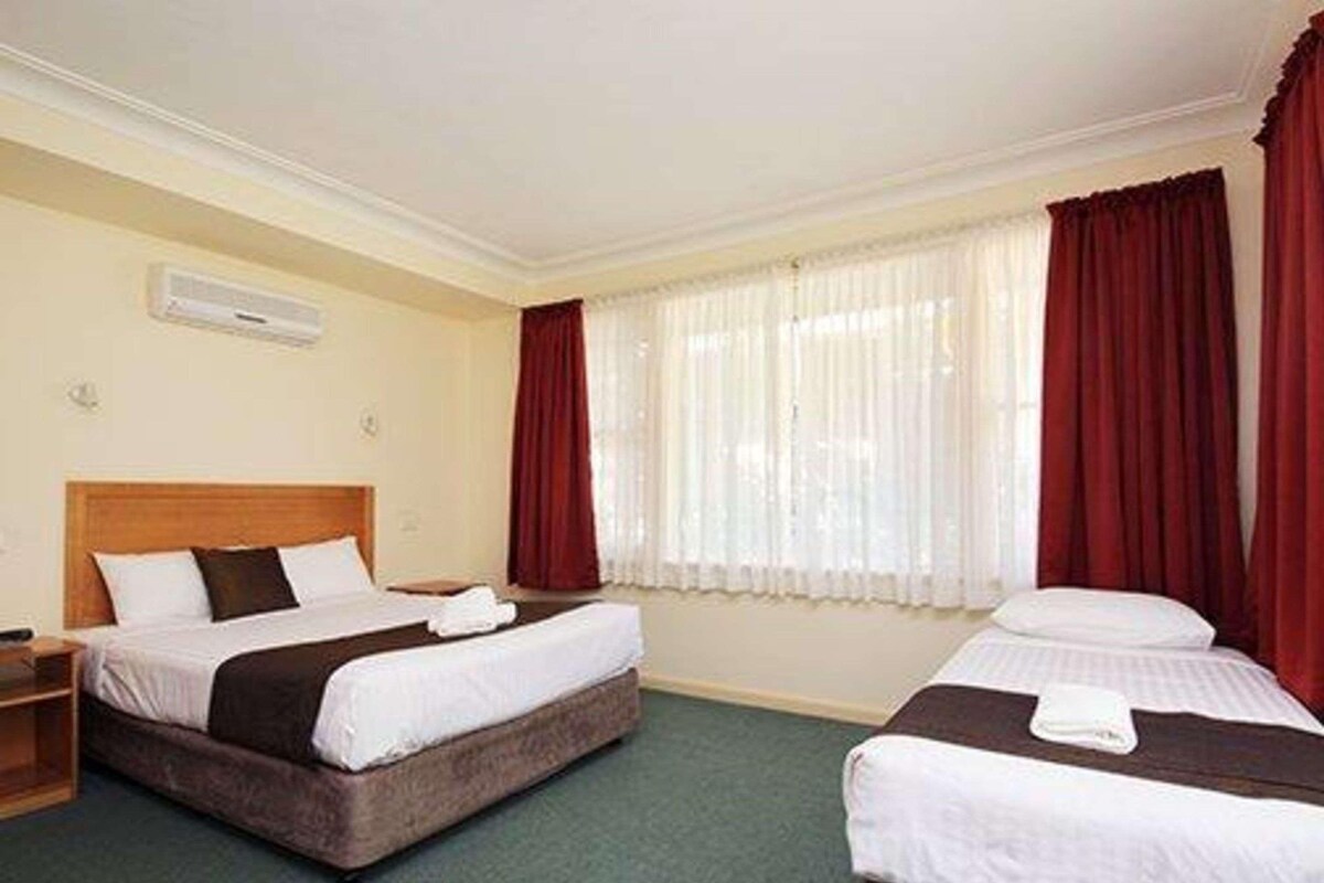 A hotel room with two beds, a wooden headboard, a window with red curtains, and a green carpet.