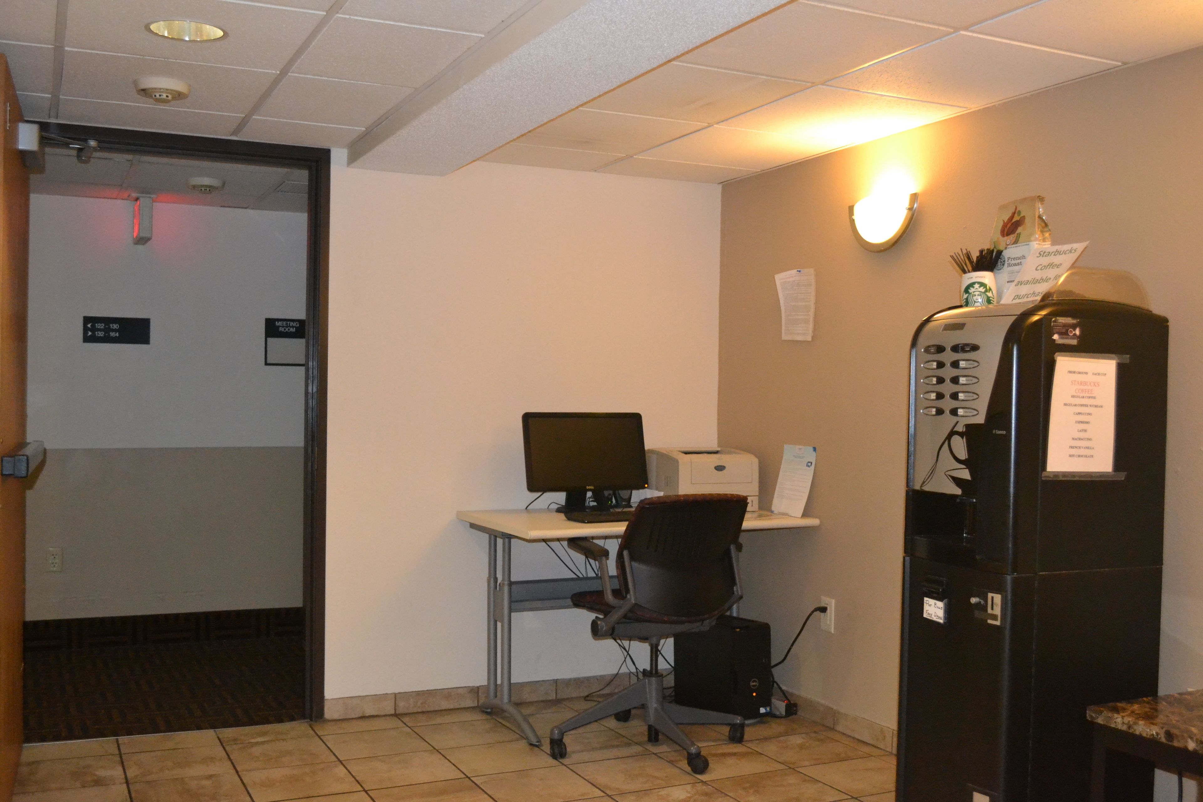 A meeting room with a desk, computer, and printer. There is a coffee machine and a wall-mounted light fixture.
