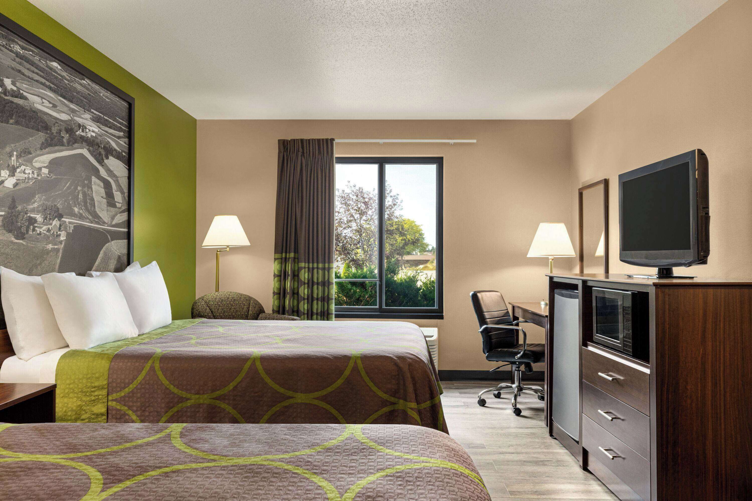 A hotel room with two beds, a desk with a television, and a window with a view of greenery.