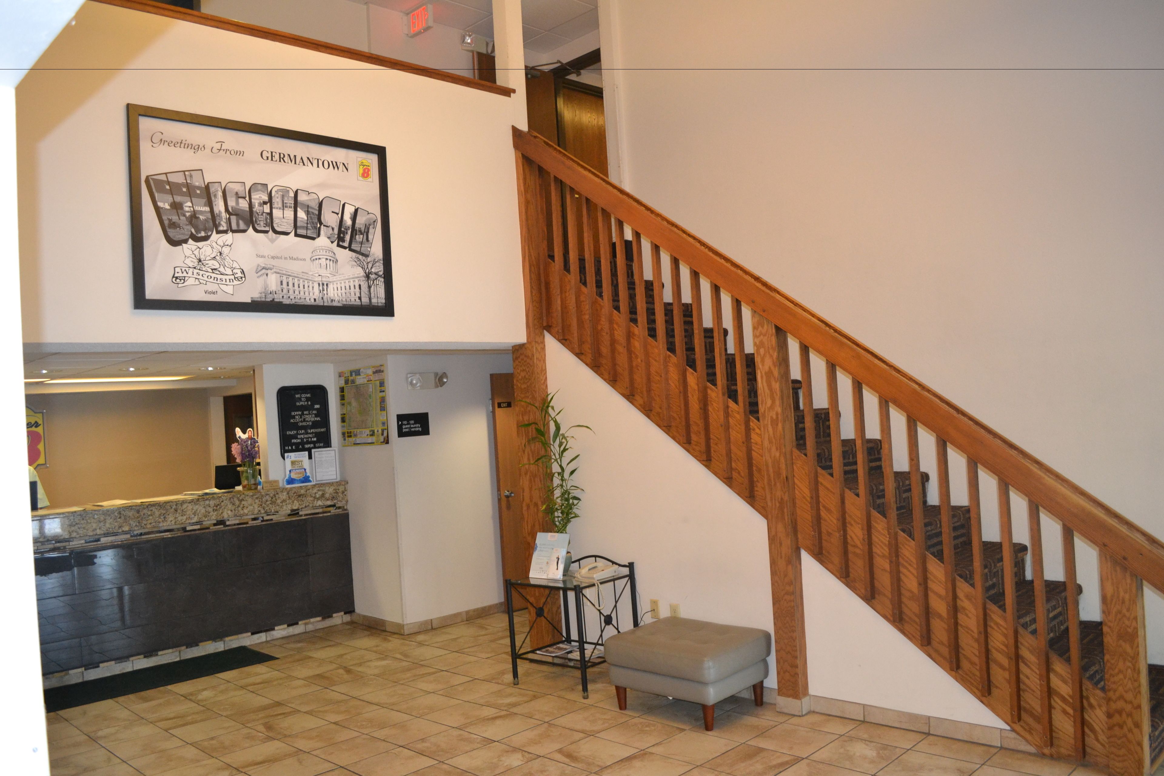 Un área de recepción con una escalera de madera, un cartel de bienvenida enmarcado, un mostrador de recepción y una pequeña mesa con una planta.