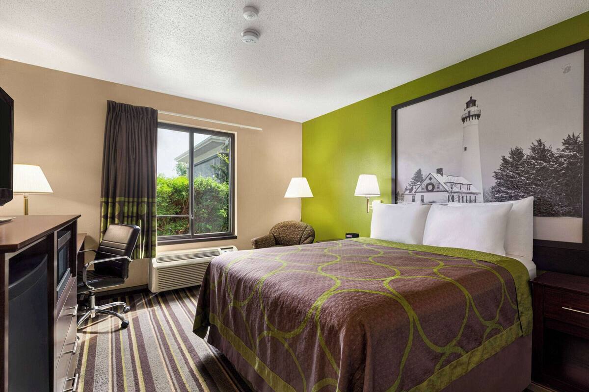 Une chambre d’hôtel avec un grand lit, deux lampes de chevet, un bureau avec une chaise, une fenêtre avec des rideaux et un tableau représentant un phare.