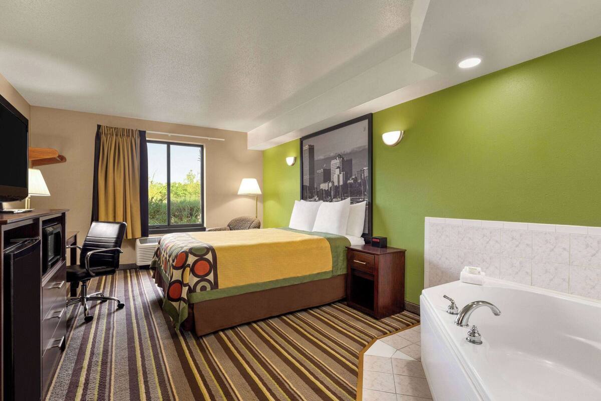 Une chambre d’hôtel comprenant un lit, un bureau avec une télévision, une chaise, une fenêtre avec des rideaux, une salle de bain avec une baignoire et un mur vert orné d’une image représentant un paysage urbain.