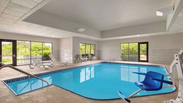Relax by the pool with lounge chairs and a sunbed, surrounded by a high ceiling and large windows offering views of the lush outdoors.