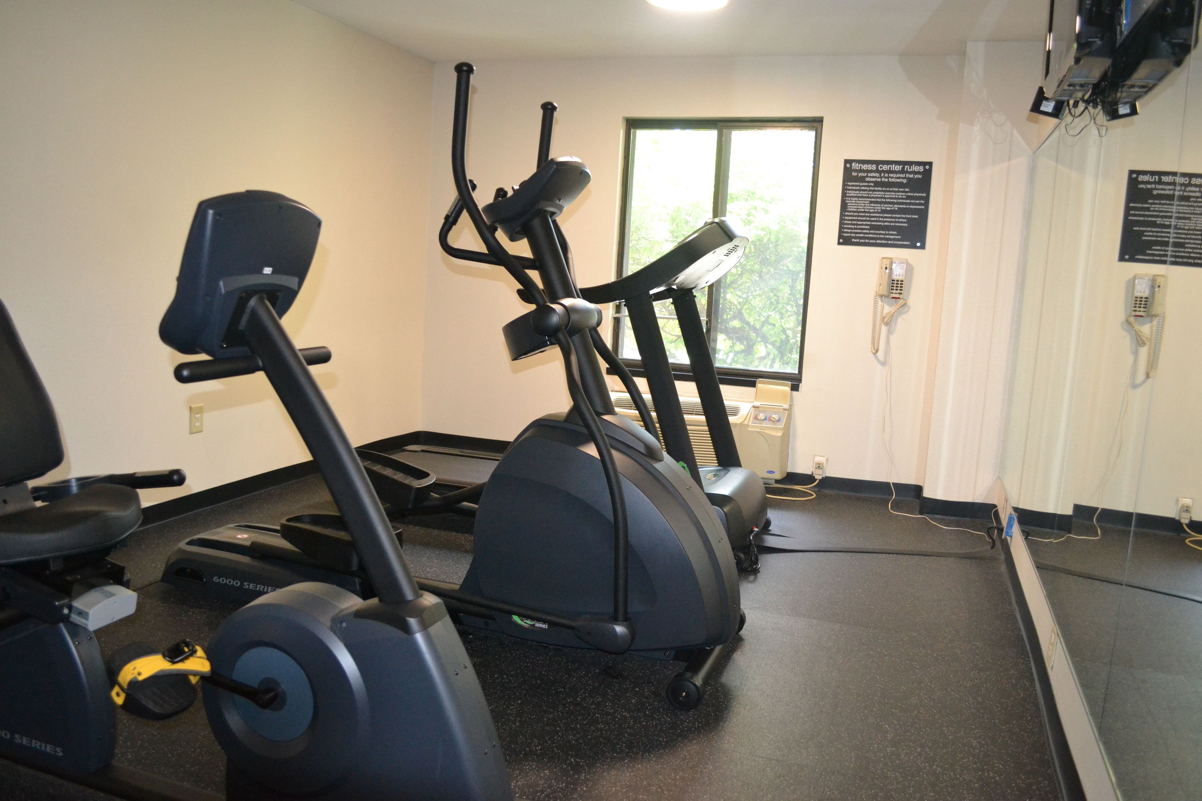 Un gimnasio con máquinas de cardio, una cinta de correr y una bicicleta estática. Hay dos carteles en la pared, uno con las normas del gimnasio y otro con un número de teléfono.
