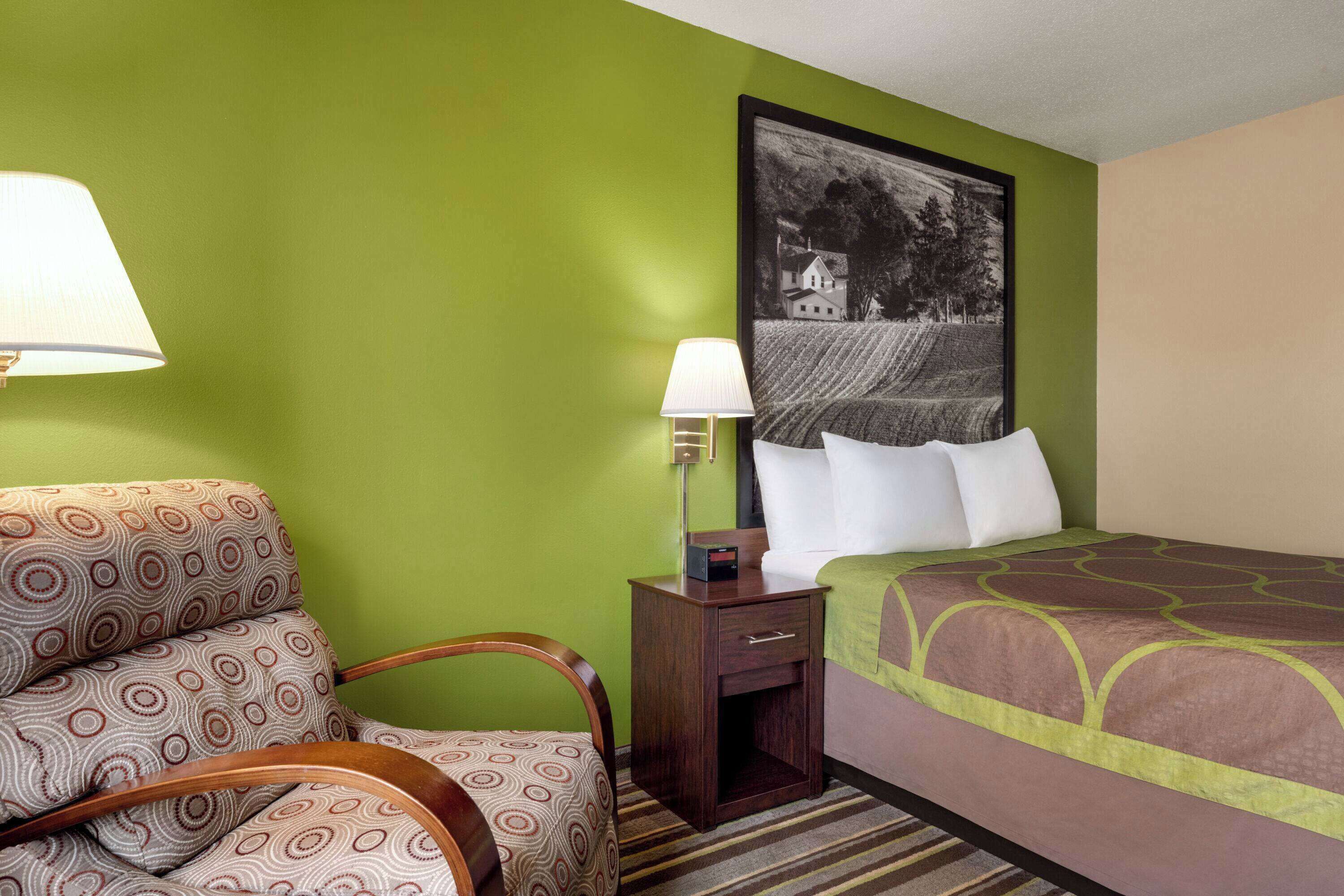 Une chambre d’hôtel avec un mur vert, un lit recouvert d’une couvre-lit à motifs, une chaise au design circulaire, une table de chevet avec une lampe et un tableau encadré accroché au mur.
