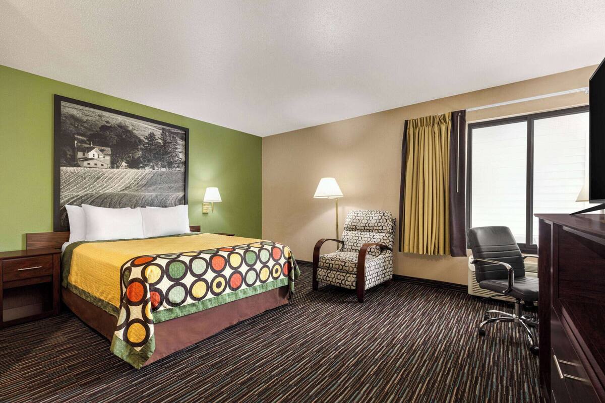 Une chambre d’hôtel avec un lit, deux chaises, un bureau, une lampe et une grande fenêtre avec des rideaux.