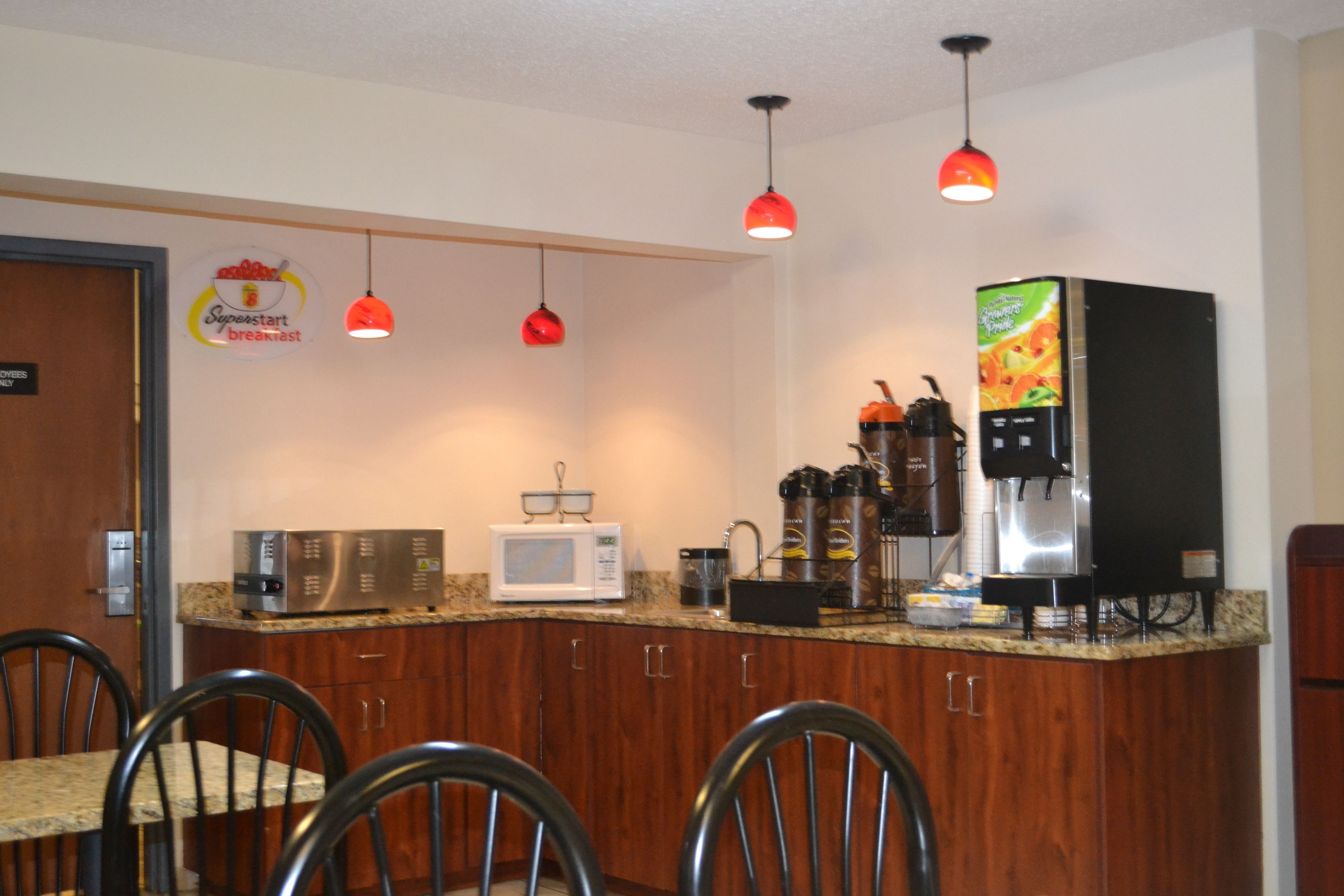 A compact kitchen with a microwave, coffee machine, and a refrigerator. There are wooden cabinets and a granite countertop. Three red pendant lights hang from the ceiling.