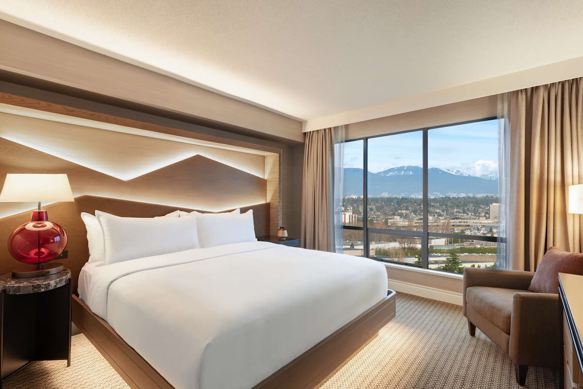 A hotel room with a large bed, a nightstand with a lamp, a chair, and a large window offering a city and mountain view.