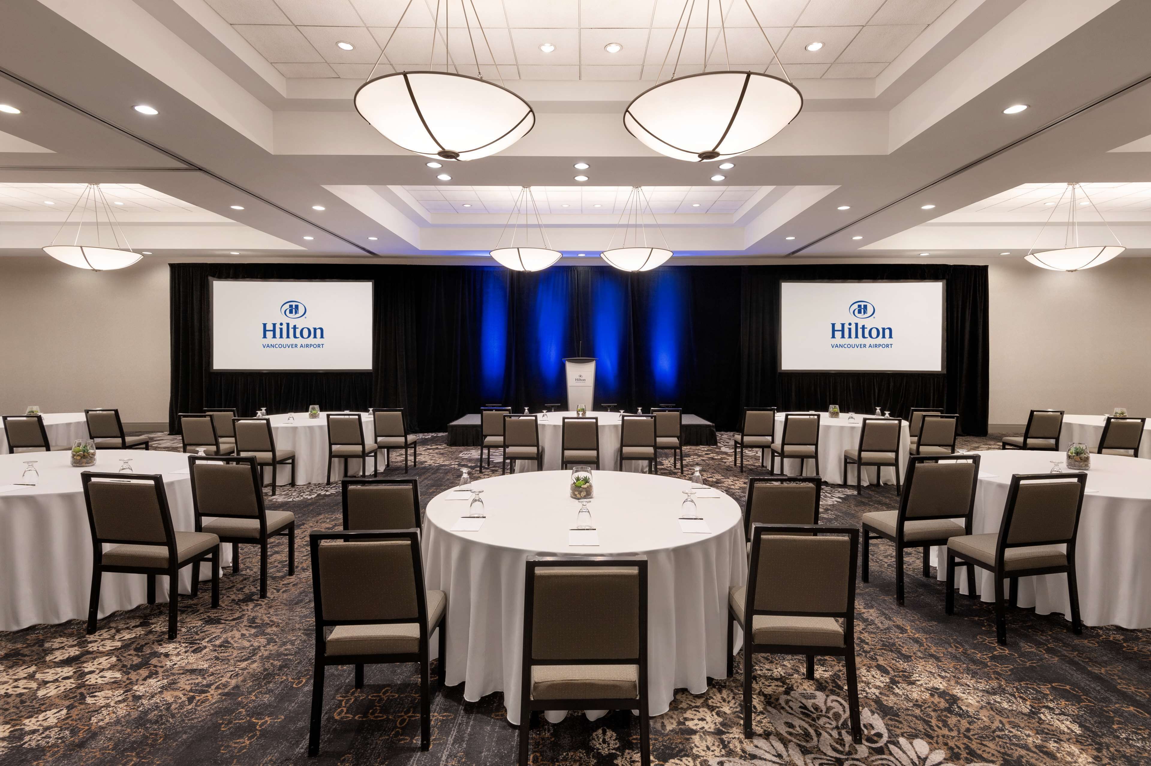 Un salón de conferencias con mesas y sillas redondas, una pantalla grande mostrando el logo de Hilton y un escenario con cortinas.