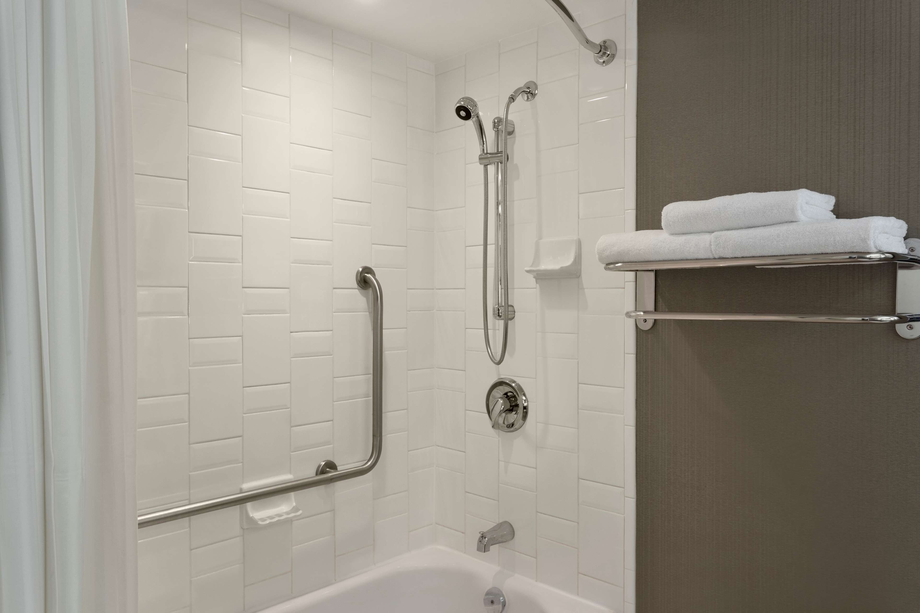A modern bathroom with white subway tile walls, a white bathtub, a shower with a handheld showerhead, and a towel rack with folded towels.