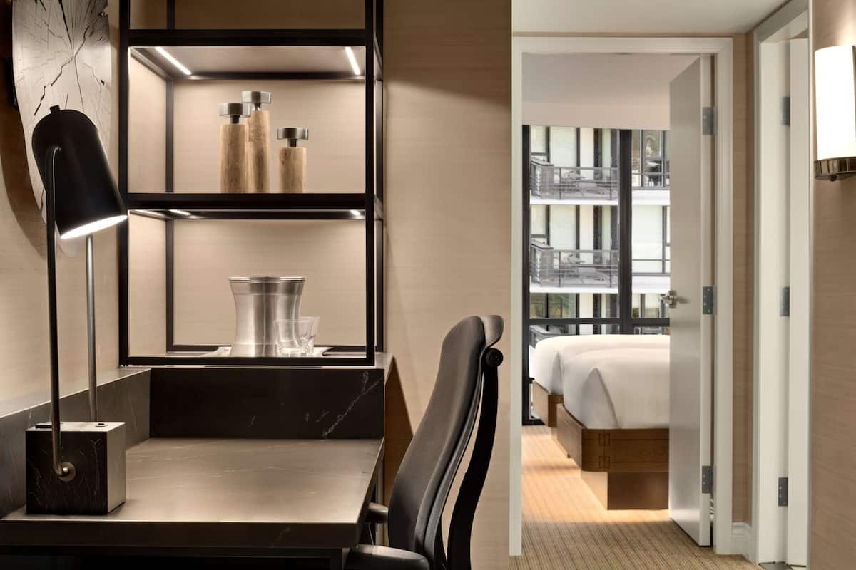A modern hotel room with a minimalist design, featuring a desk with a lamp, a chair, a bed with a wooden headboard, and a large window offering a view of the city.