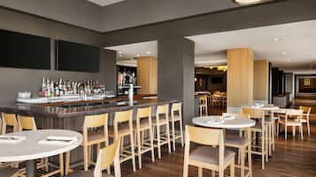 Un bar moderno con una barra larga, varios taburetes y una amplia variedad de botellas y vasos. Hay varias mesas redondas con sillas dispuestas alrededor del bar.