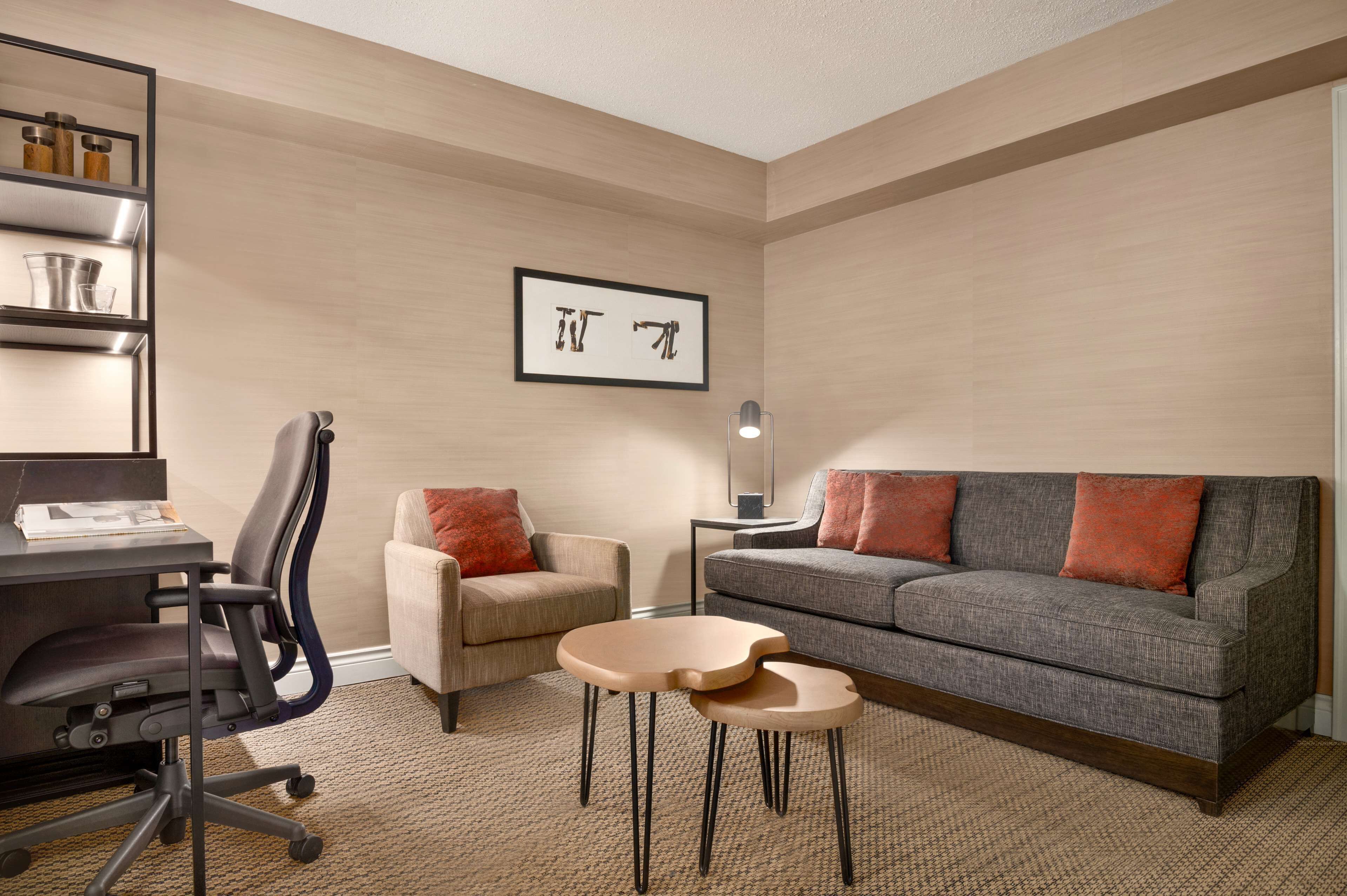 A modern living room with a grey sofa, a beige armchair, a small table, and a desk with a chair. There is a framed artwork on the wall and a lamp on a side table.