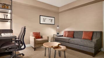A modern living room with a grey sofa, a beige armchair, a small table, and a desk with a chair. There is a framed artwork on the wall and a lamp on a side table.