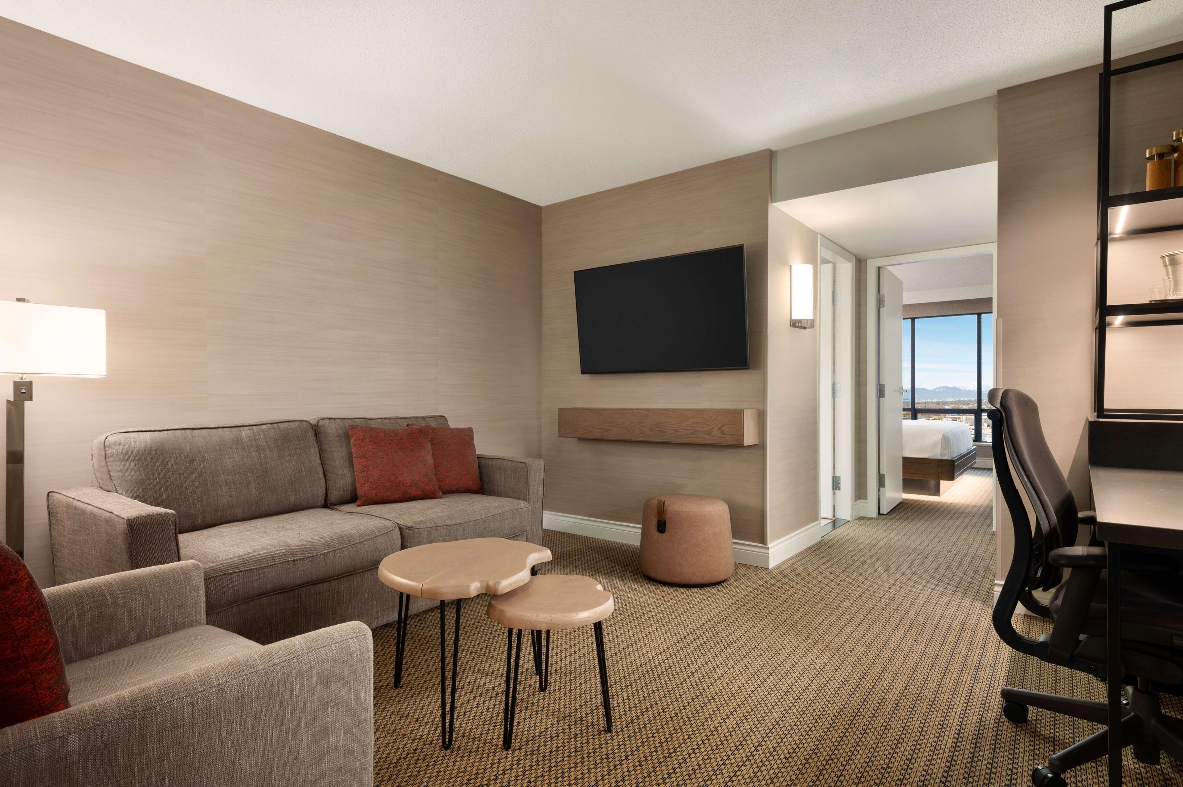Habitación de hotel con sofá, dos mesitas, televisión montada en la pared, escritorio con silla y vistas a la ciudad desde la ventana.