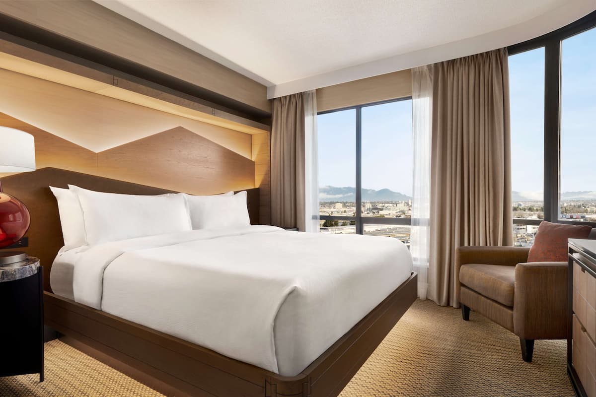 A hotel room with a large bed, a desk, a chair, and a view of the city through large windows.