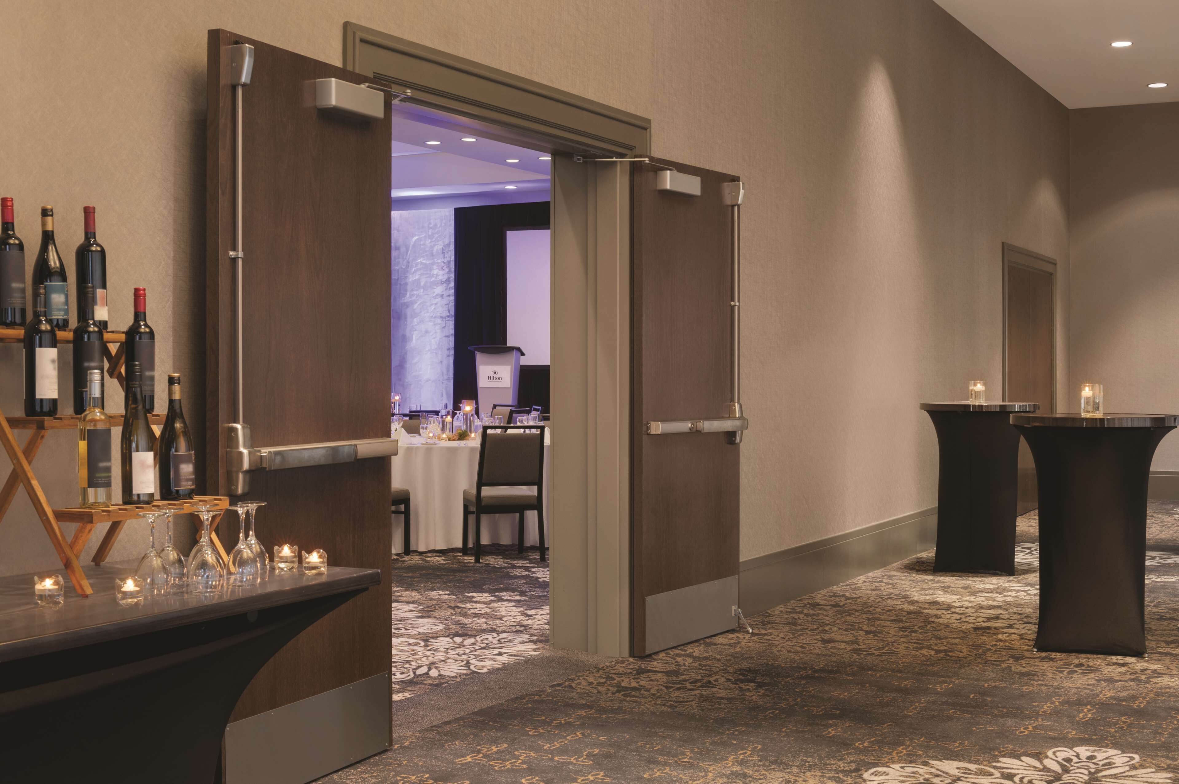 Un vestíbulo de hotel con una recepción, una zona de bar con botellas de vino y una sala de conferencias al fondo.