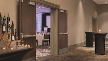 Un vestíbulo de hotel con una recepción, una zona de bar con botellas de vino y una sala de conferencias al fondo.