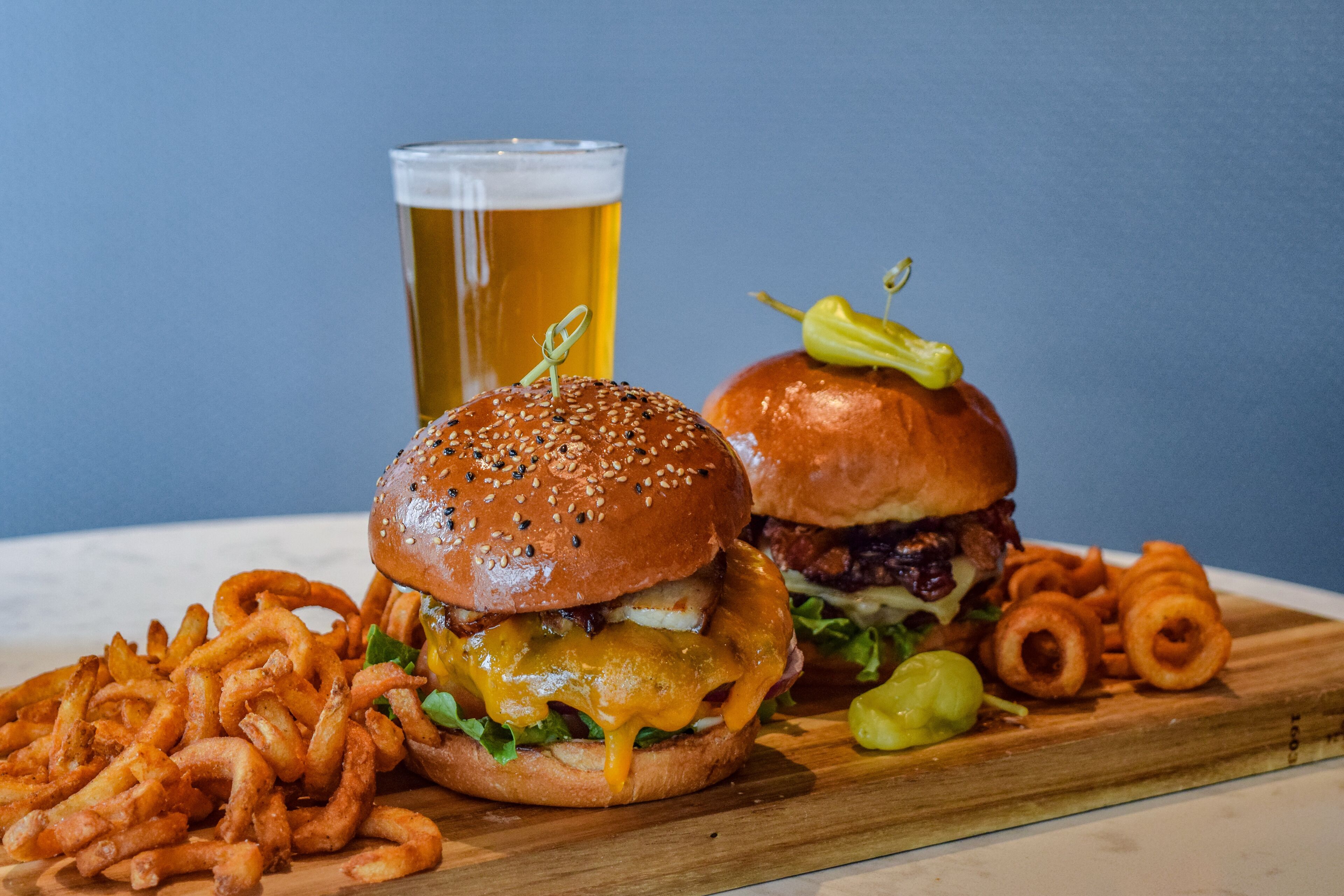 A meal with two burgers, a glass of beer, and onion rings on a wooden board.