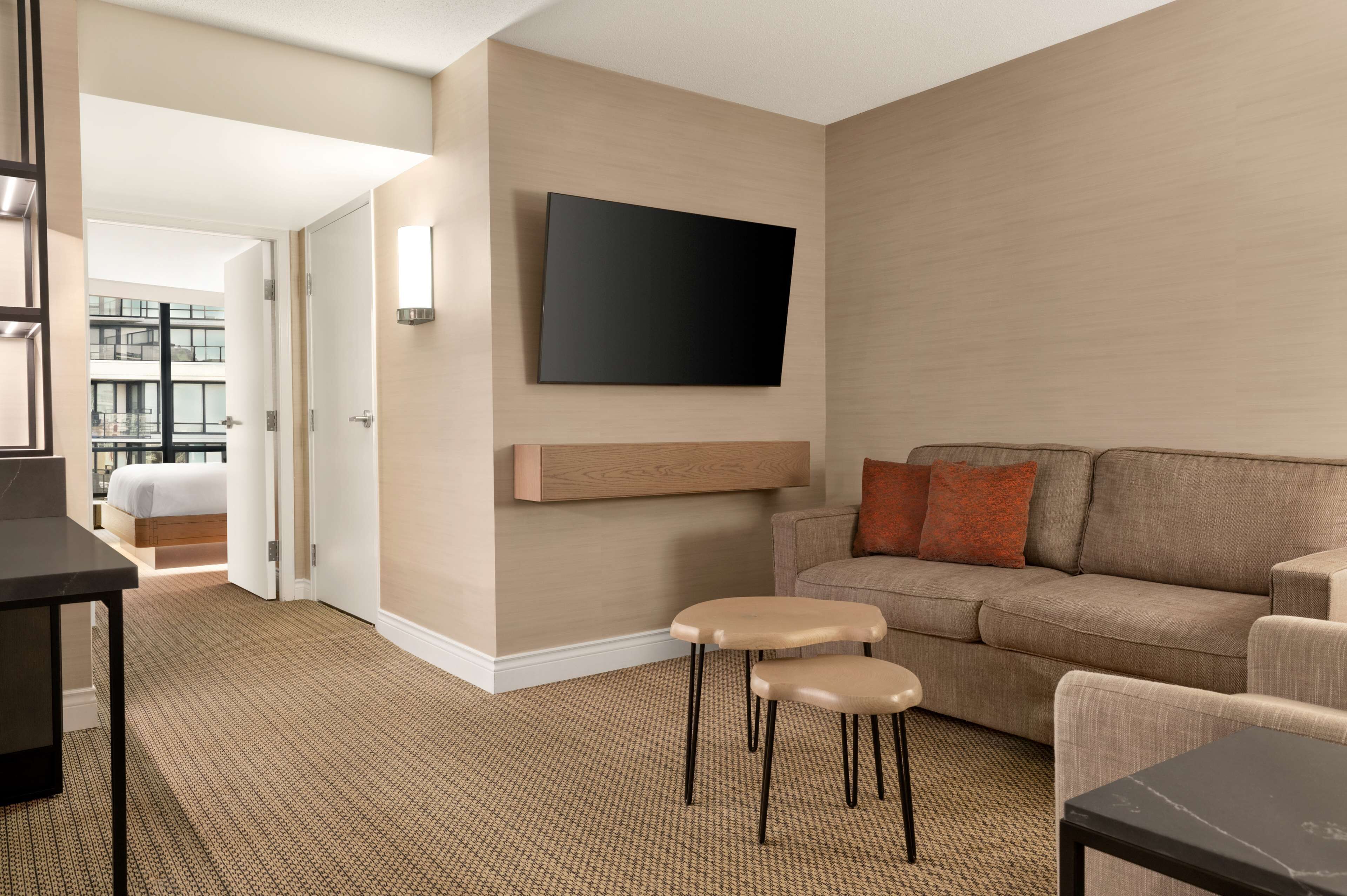 A modern hotel room with a large flat-screen TV mounted on the wall, a sofa, two small stools, and a bed with a view of the cityscape.
