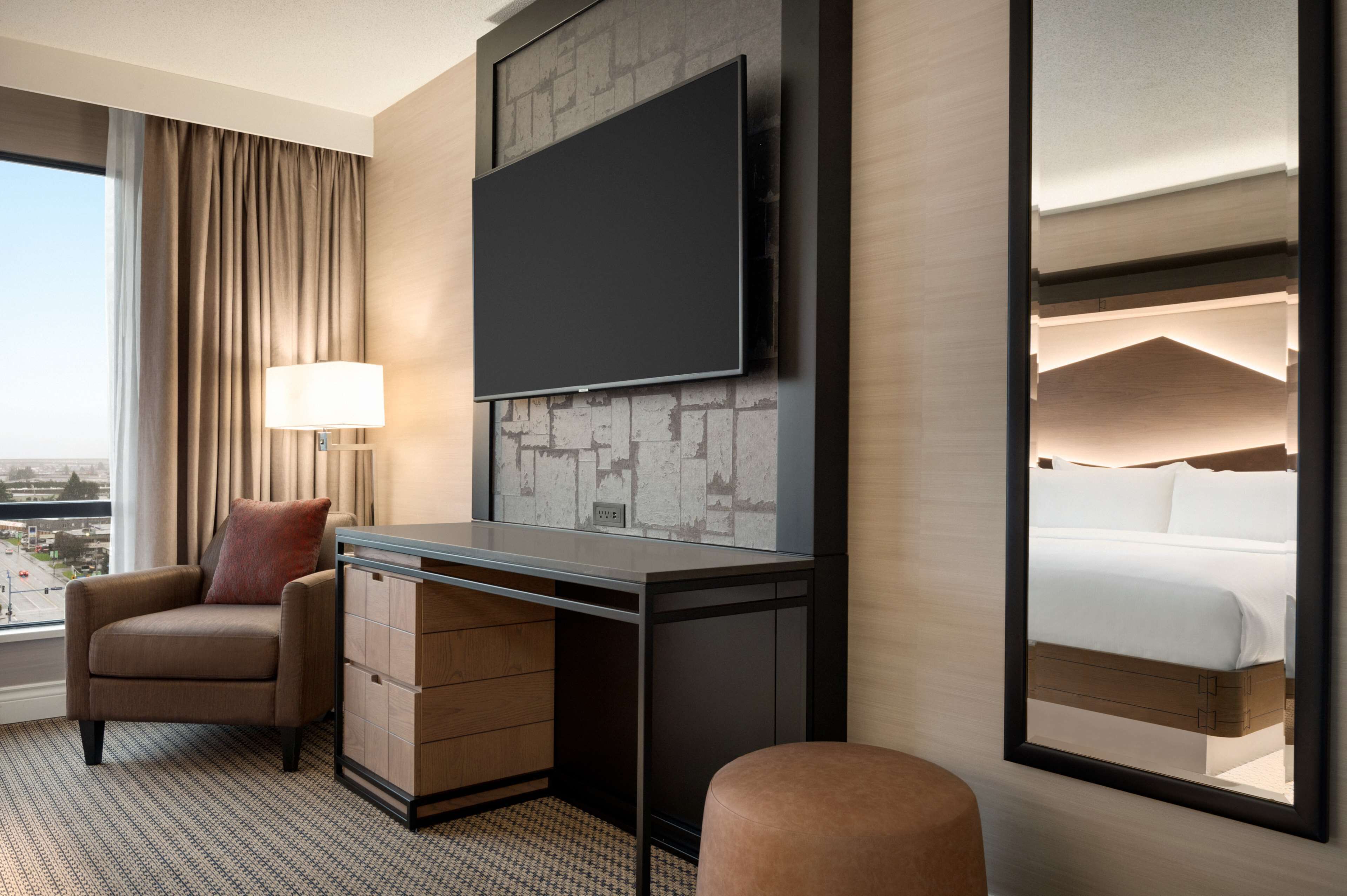 A hotel room with a large flat-screen TV mounted on a brick-patterned wall, a desk with a lamp, a comfortable armchair, and a view of the city through a large window.
