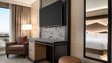 A hotel room with a large flat-screen TV mounted on a brick-patterned wall, a desk with a lamp, a comfortable armchair, and a view of the city through a large window.