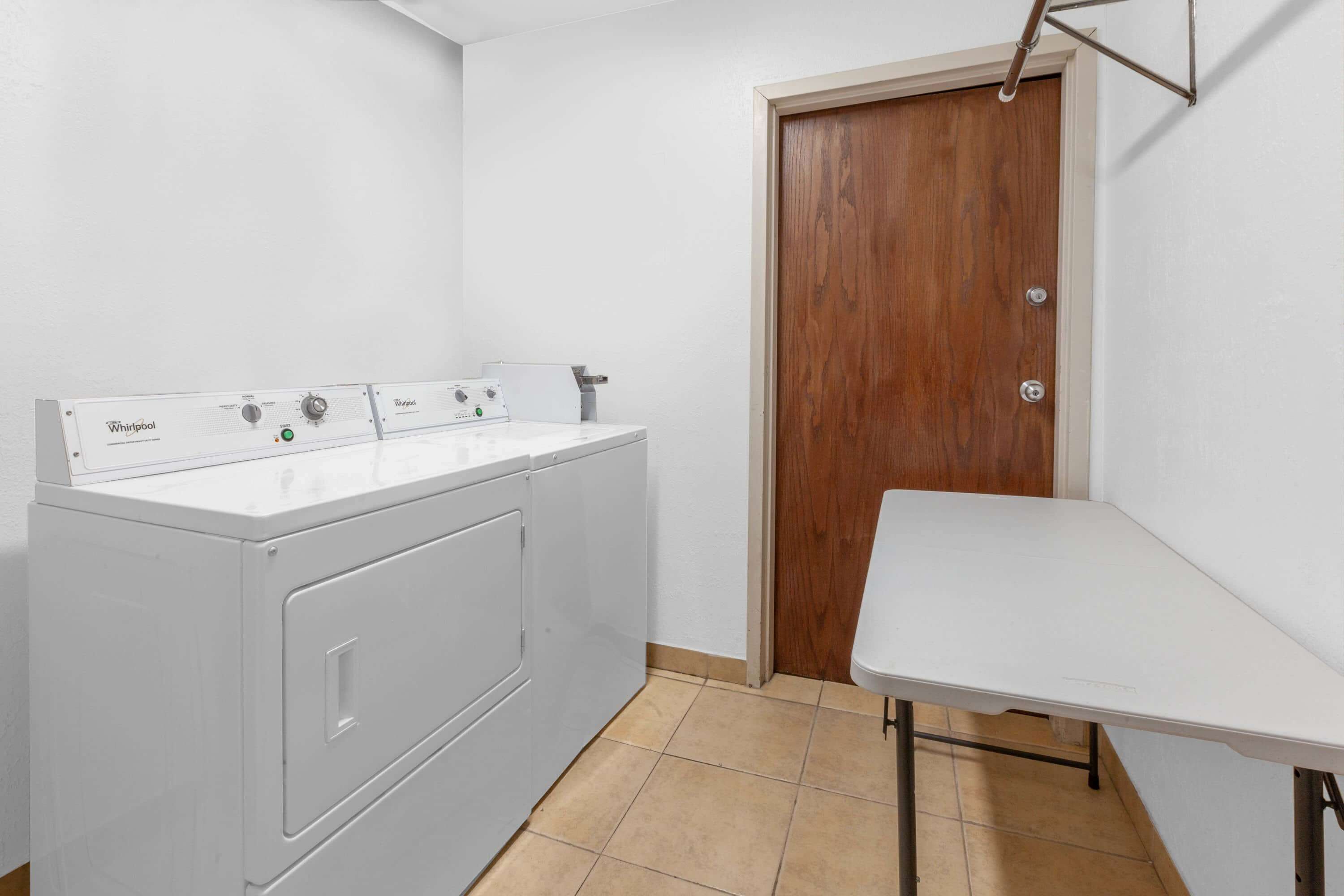 A laundry area with a white Whirlpool washer and dryer, a wooden door, and a white table.