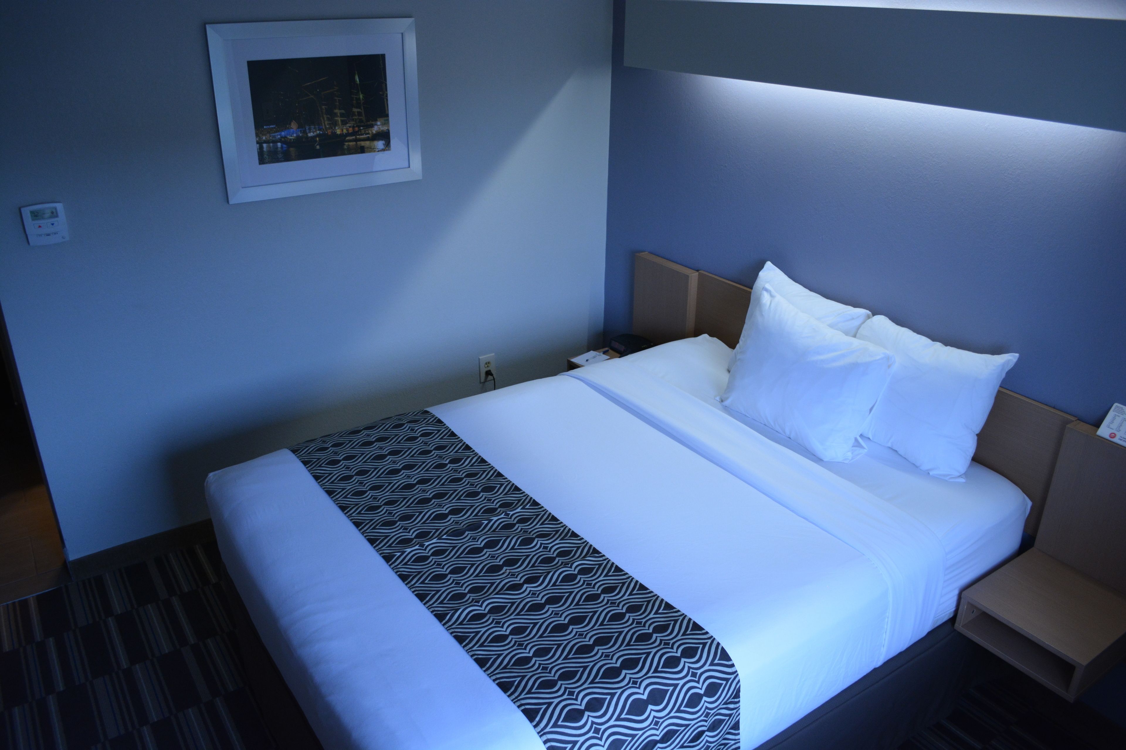 A hotel room with a neatly made bed, a framed picture on the wall, and a bedside table with a lamp.