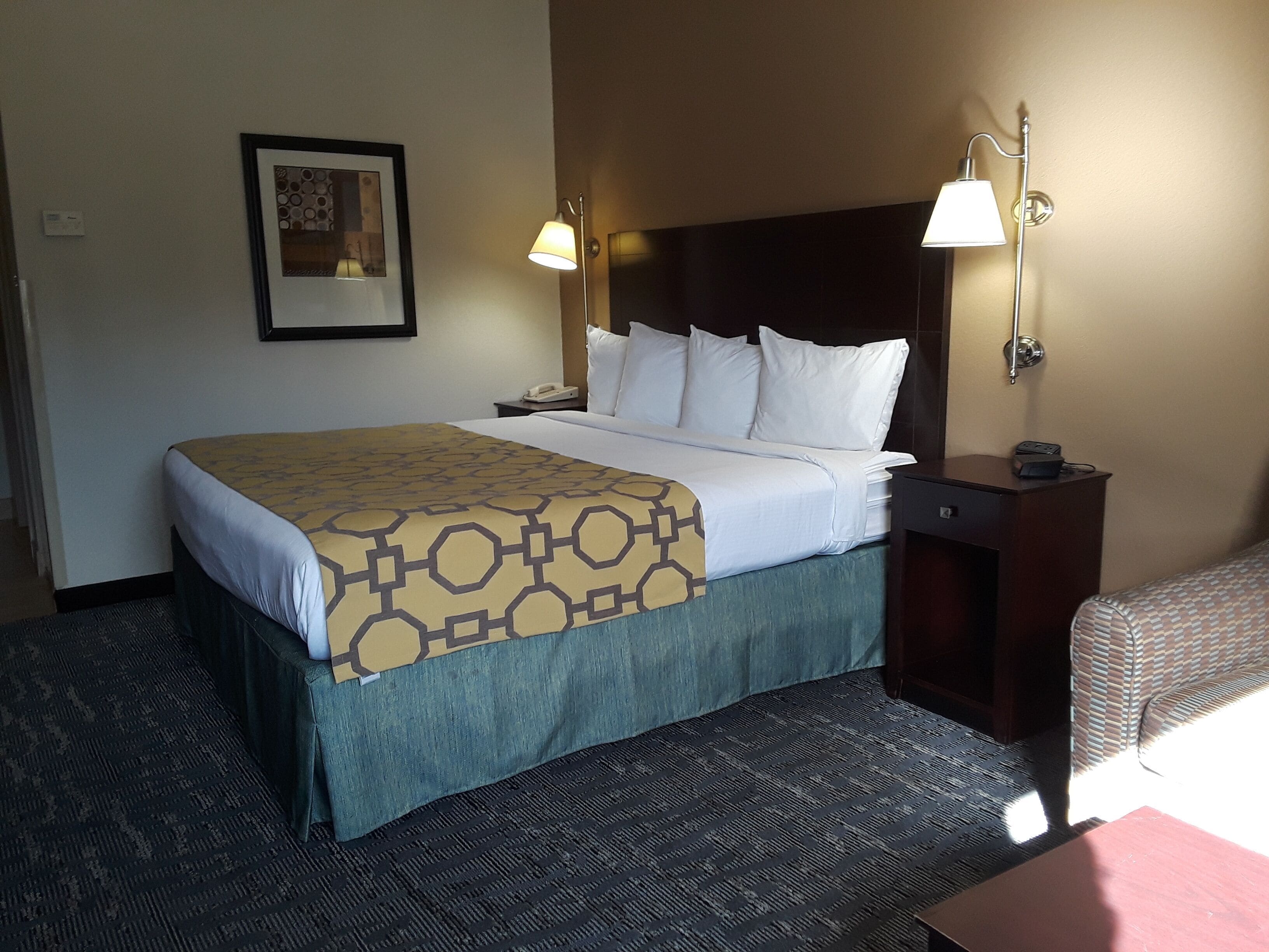 A hotel room with a bed, two bedside lamps, a nightstand with a phone, a framed picture on the wall, and a patterned carpet.